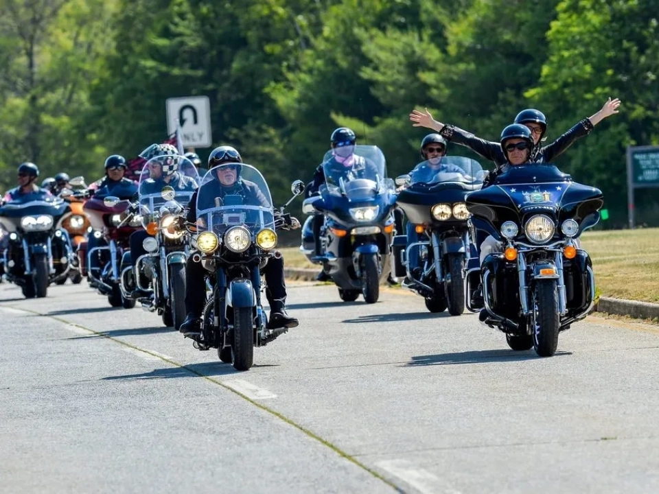 A group of motorcyclists with custom motorcycle seats riding on a road, with the lead riders raising their hands joyfully. All are wearing helmets.